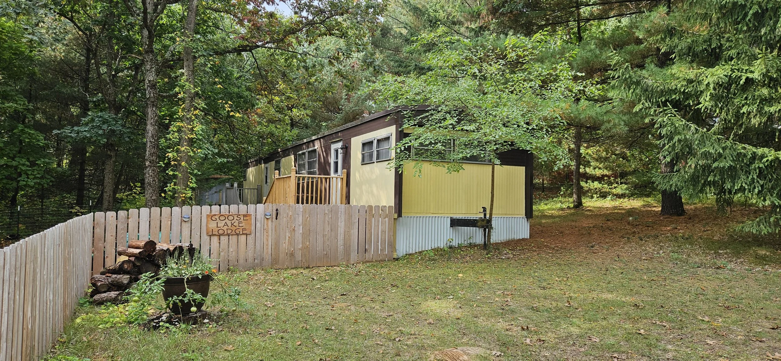 A mobile home with yellow and brown siding situated in a wooded area surrounded by green trees and a grassy yard, enclosed by a wooden fence with a sign that reads 'Goose Lake Lodge.' There is a woodpile in a planter and a grassy area in front of the home.