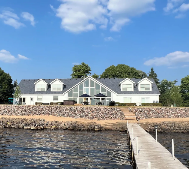 A large white lakeside house with a dark gray roof, overlooking a dock on the water with a stone retaining wall and outdoor seating area.