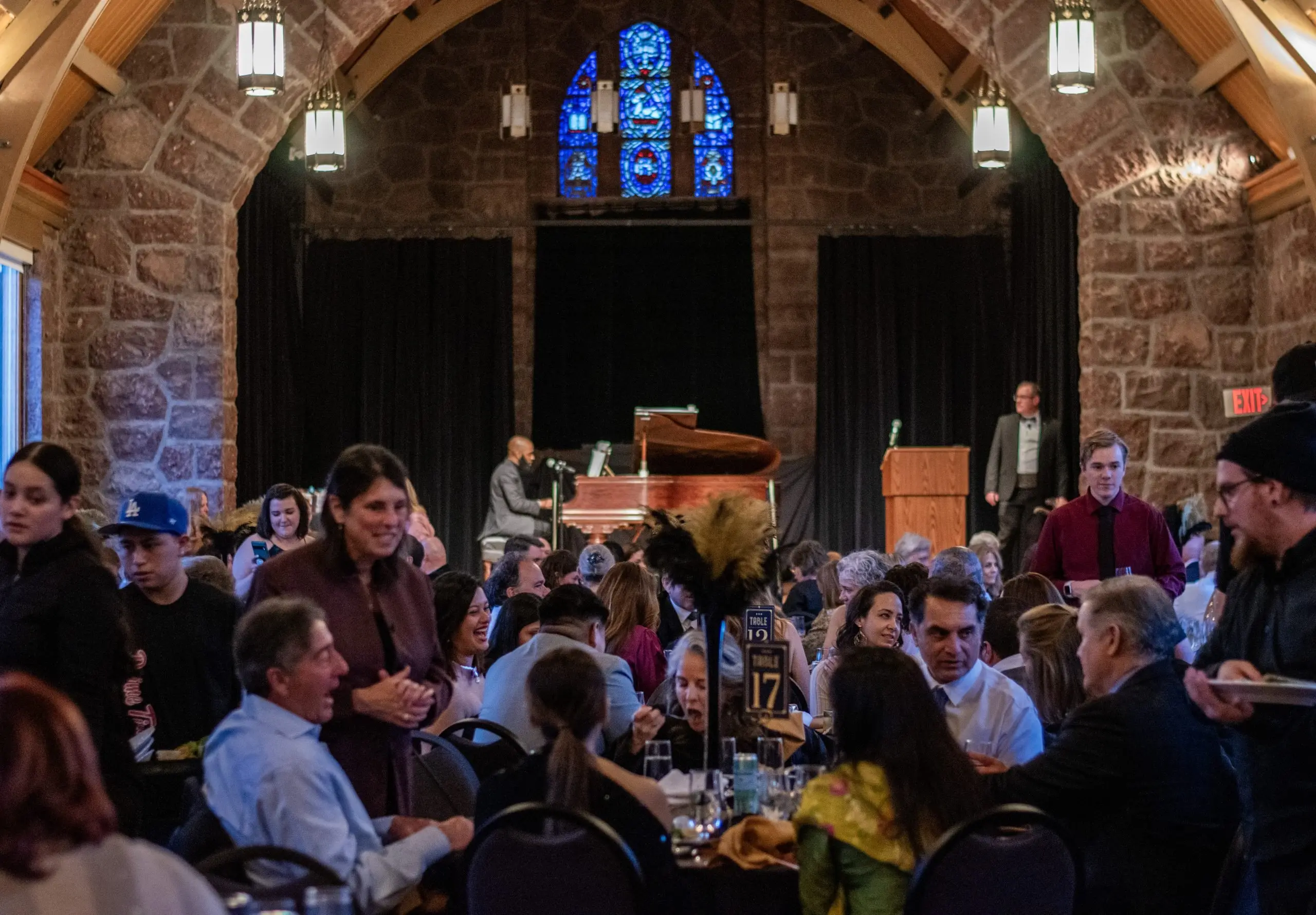 Wide view of a stone hall with a gothic timber arch, blue stained-glass window and brass pendant lanterns, set with round tables and guests.