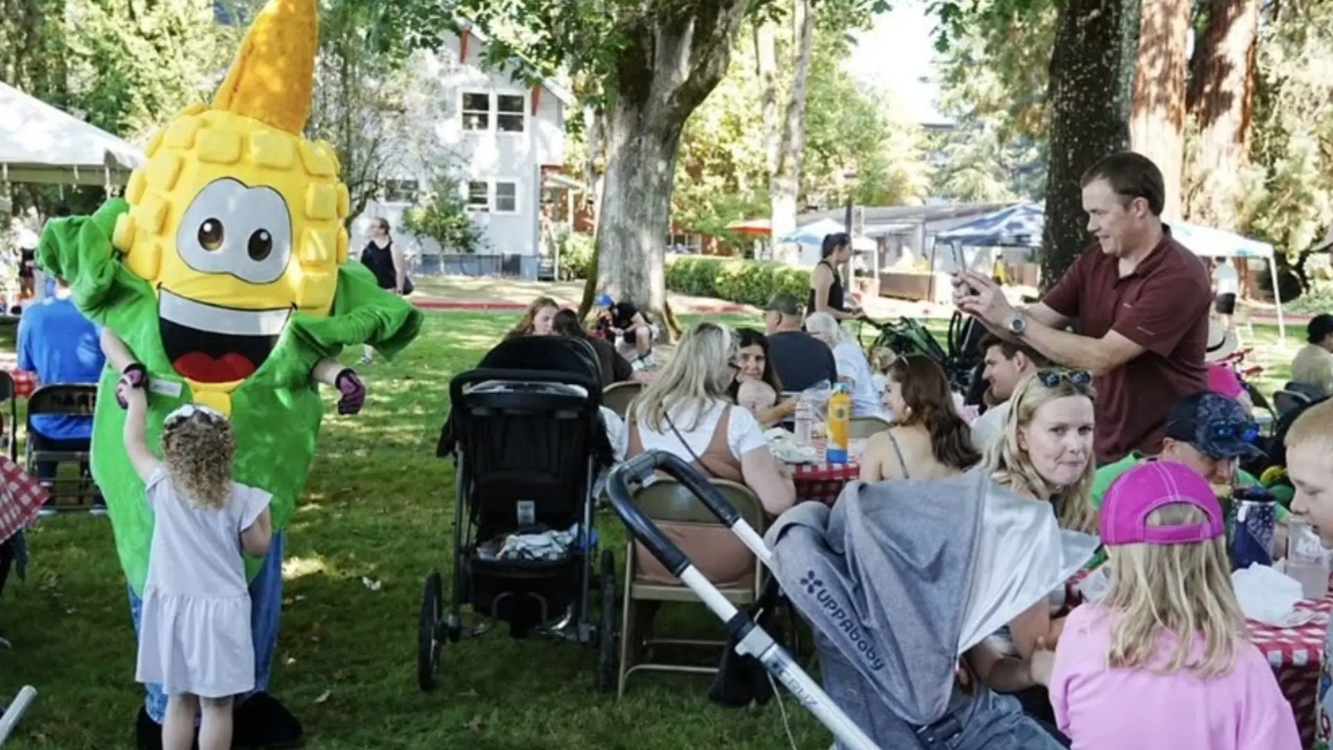 A child high-fives a smiling corn mascot as families gather around picnic tables at an outdoor community event