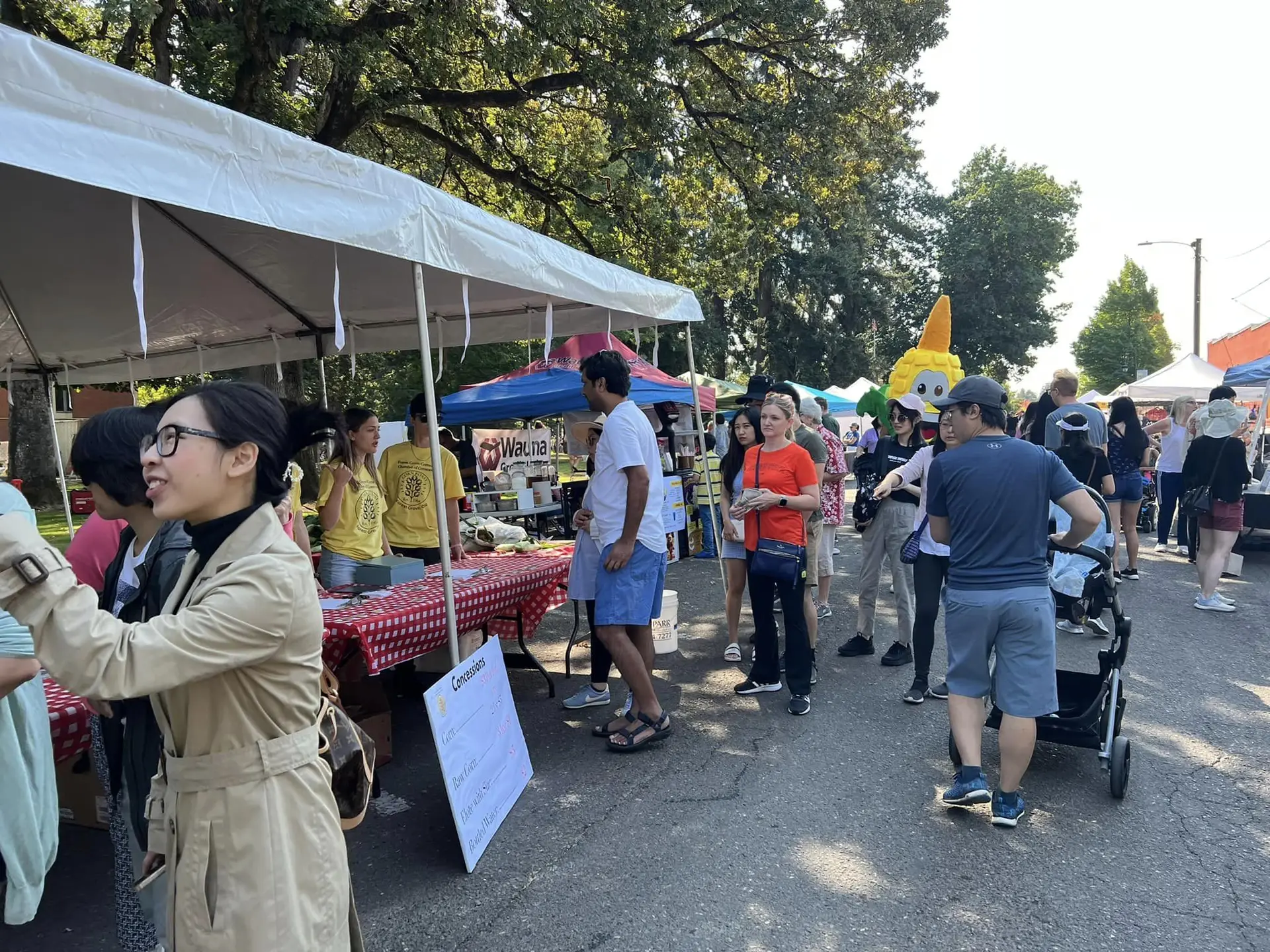 Rows of white canopy tents down a closed street under mature trees, families walking between vendor tables, a corn-cob mascot in the crowd.