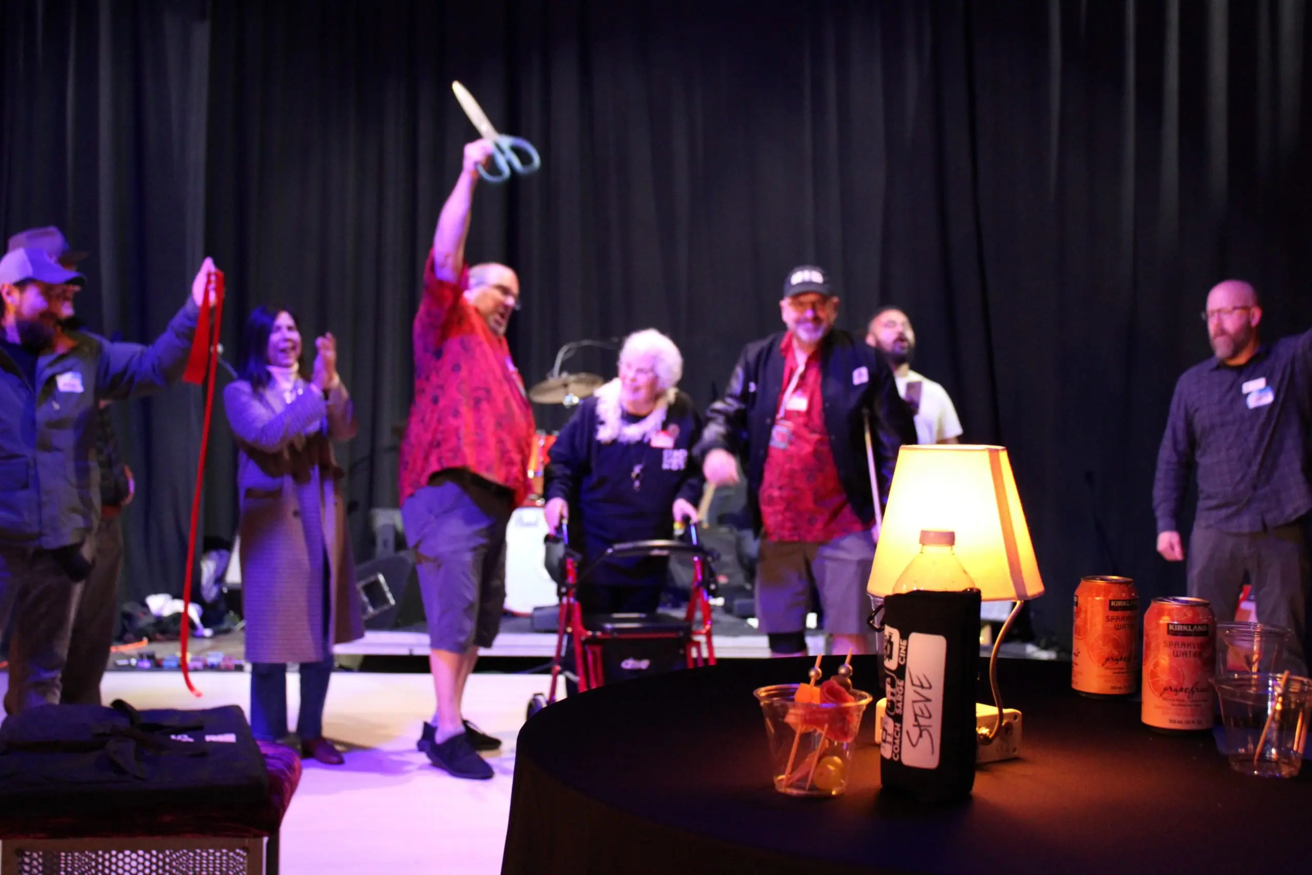 A person holding oversized ceremonial scissors on a small stage in front of a dark curtain.