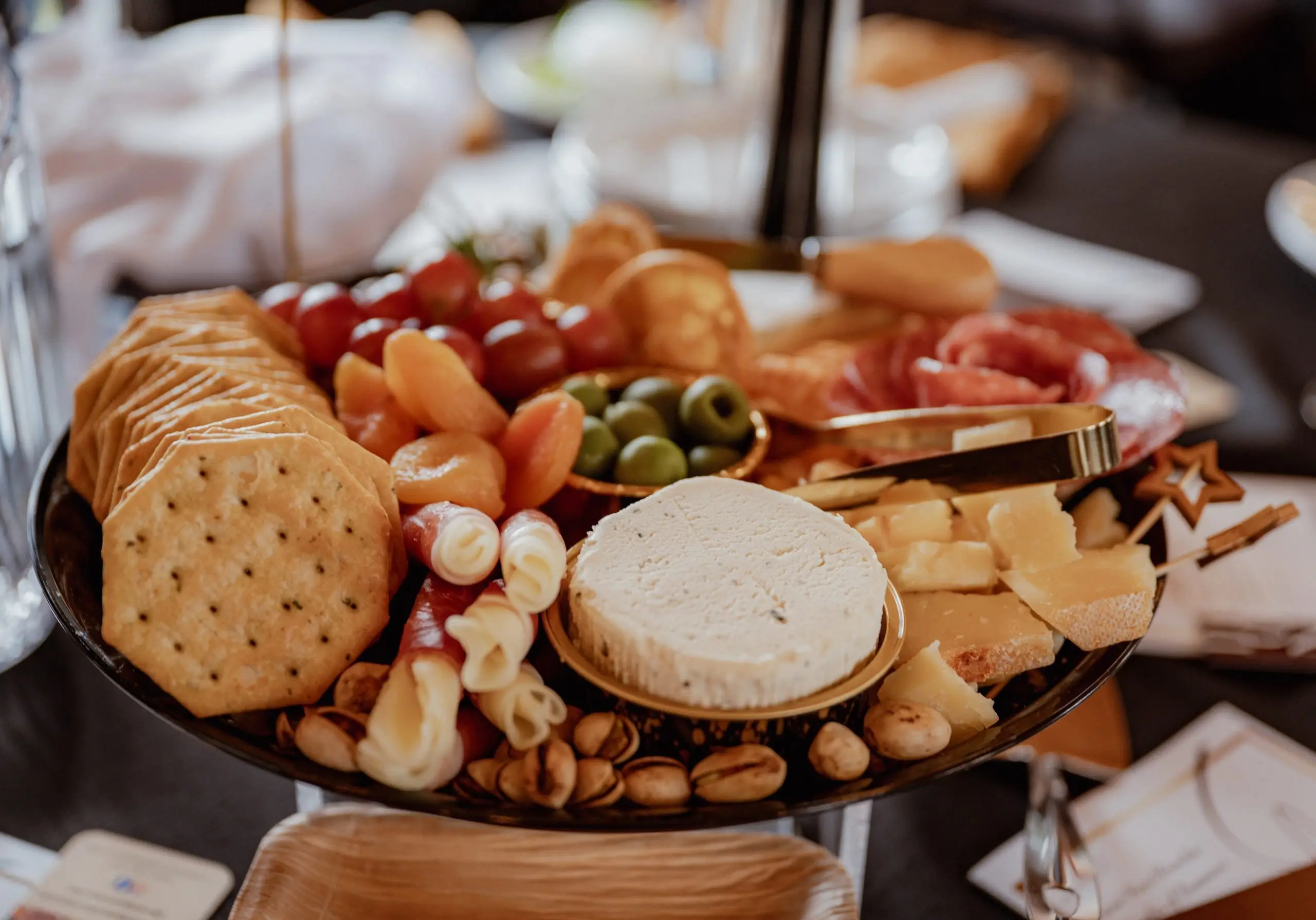A laden charcuterie board shot at a shallow angle: a soft cheese round, sliced cheeses, crackers, breadsticks, olives, grapes and cured meats.