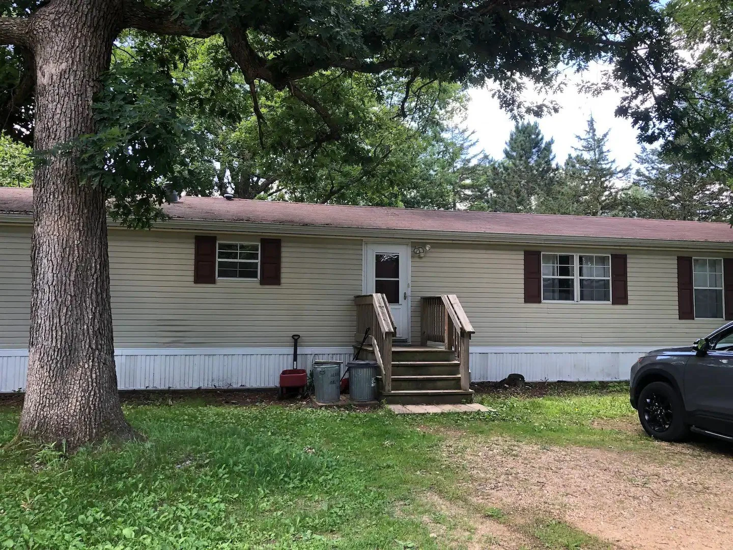 A single-story mobile home with beige siding and dark red shutters, set in a green yard with mature trees. A small wooden staircase leads to a back door, and there are trash cans near the stairs. A black vehicle is parked to the right, and the driveway is a mix of gravel and dirt.