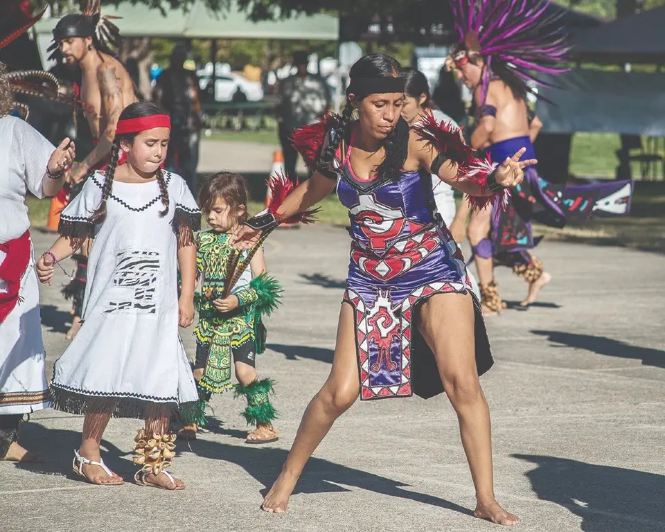 Danza Azteca dancers on a paved area in a park: a woman mid-step in purple and red regalia, a girl in a white embroidered dress and a toddler in green feathered regalia.