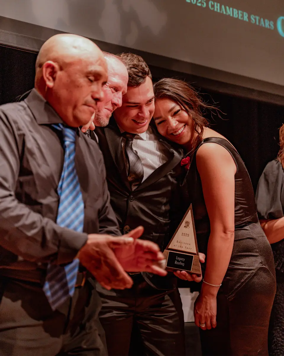 A smiling group celebrating together on stage, one woman leaning in beside a man holding a 2025 Business of the Year award, fellow recipients gathered around them