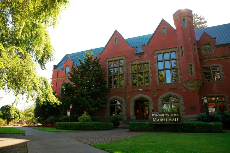 Marsh Hall at Pacific University, a red brick building with steep gables and a blue metal roof, a dark wooden sign reading Welcome to Pacific Marsh Hall in front of it.