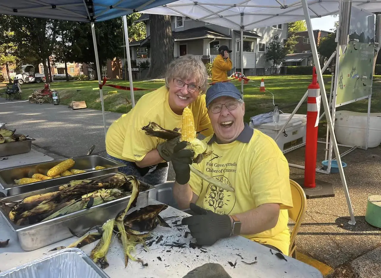 Two volunteers in yellow chamber T-shirts laughing at the camera behind trays of roasted corn, one holding a shucked cob in a heavy work glove.