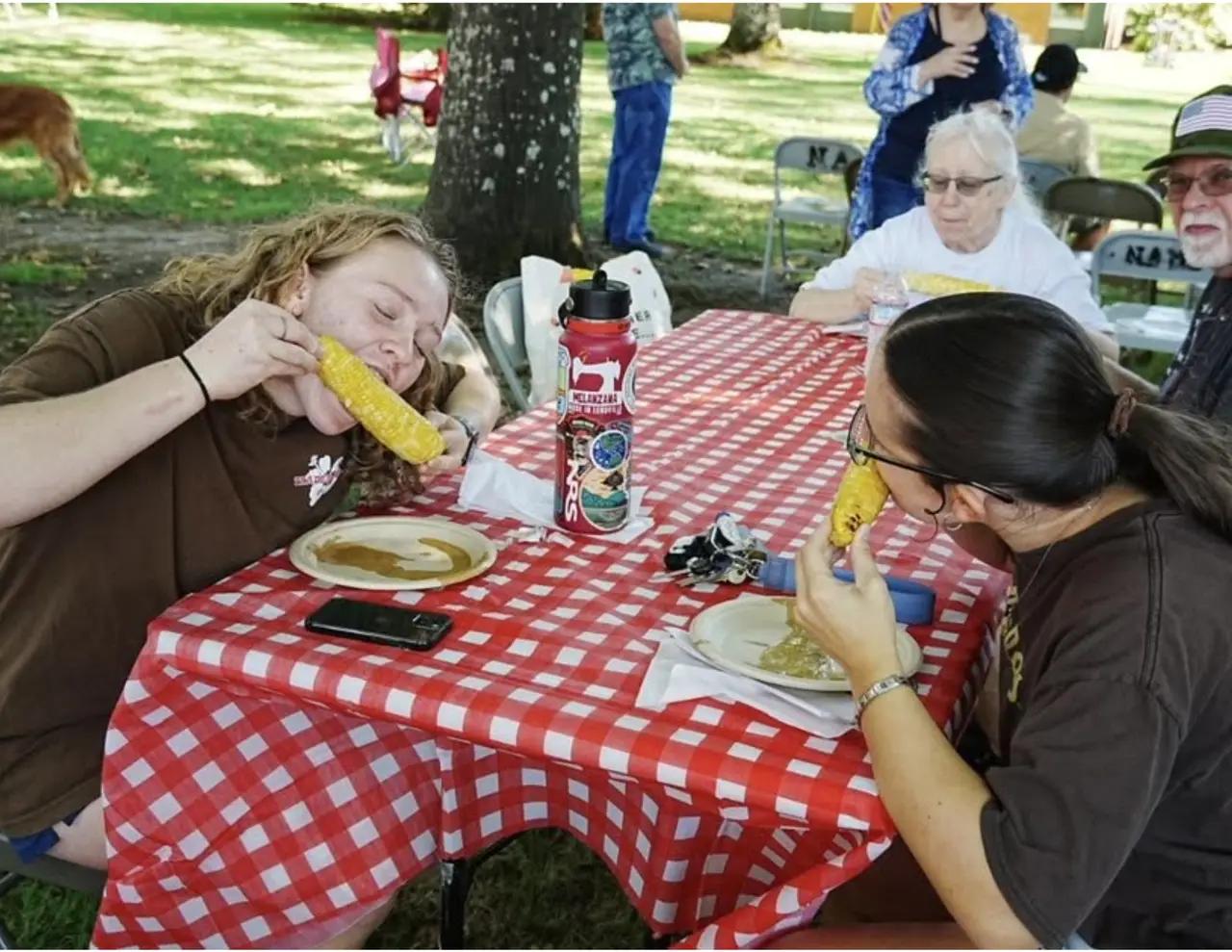 A woman biting into an ear of roasted corn at a red-and-white checked picnic table, a second woman beside her.