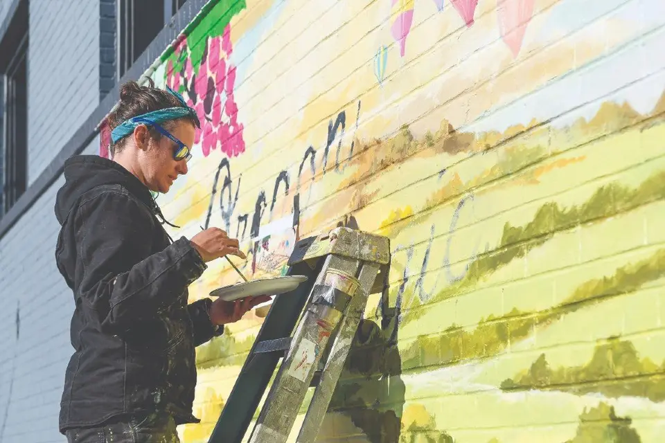A muralist on a paint-spattered ladder brushing detail into a large pastel mural on a downtown wall, holding a palette.