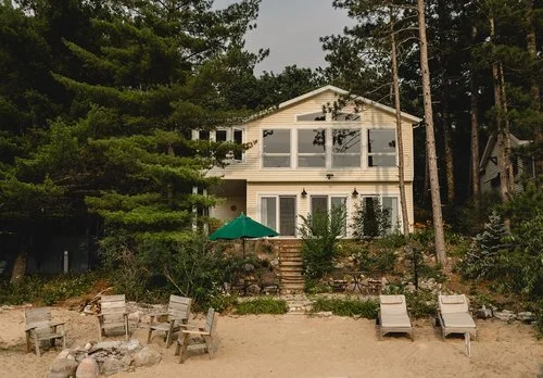 Beach house surrounded by trees with outdoor seating and a sandy beach in the foreground.
