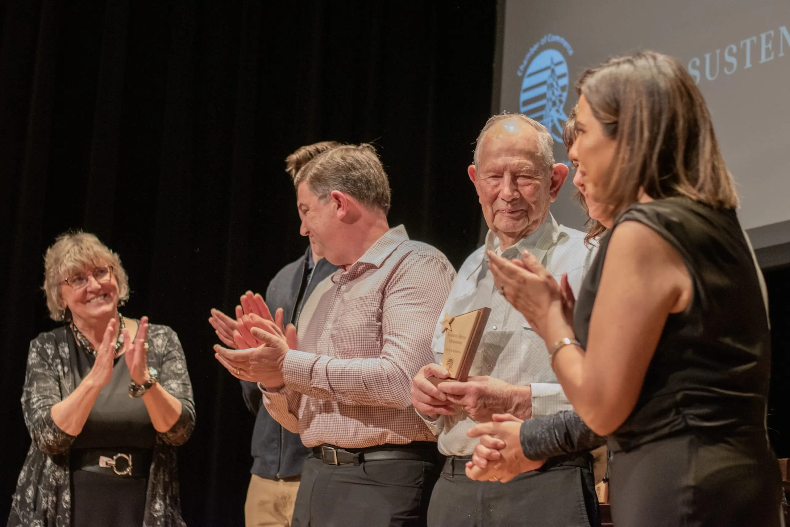 An older man holding a wooden award on stage while four people around him applaud.