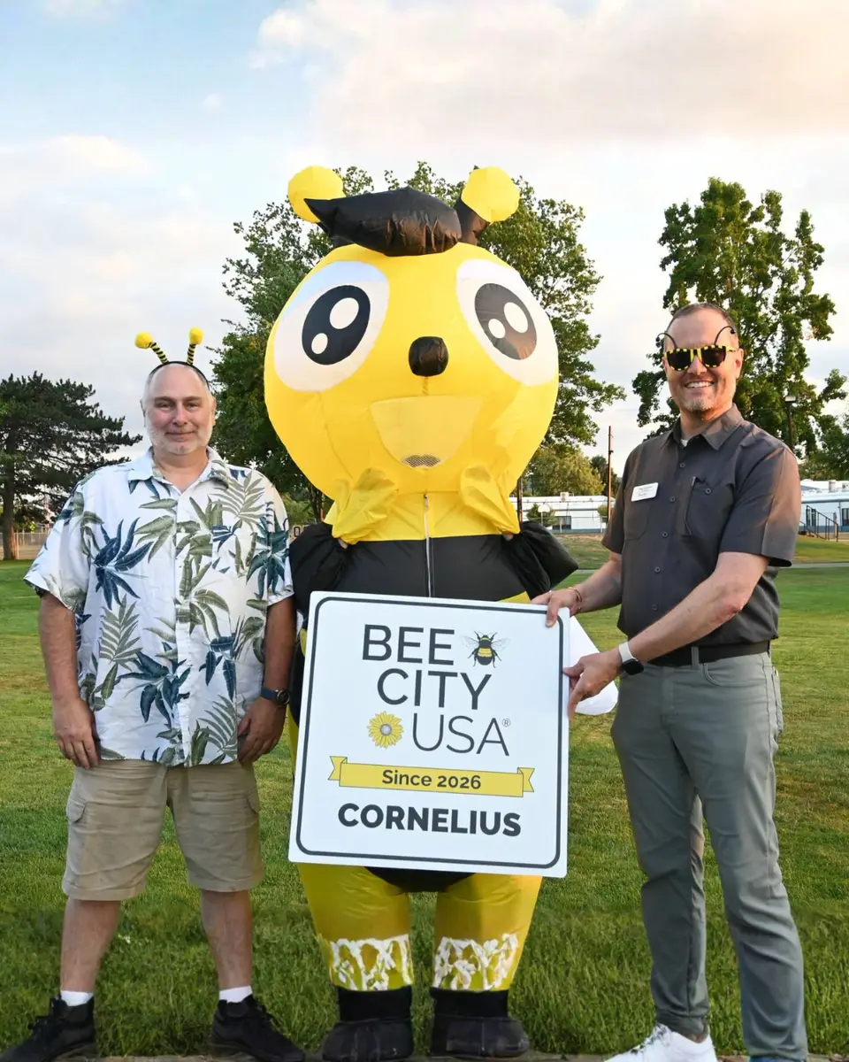 Two men pose with a large bee mascot holding a Bee City USA sign celebrating Cornelius becoming a designated Bee City in 2026.