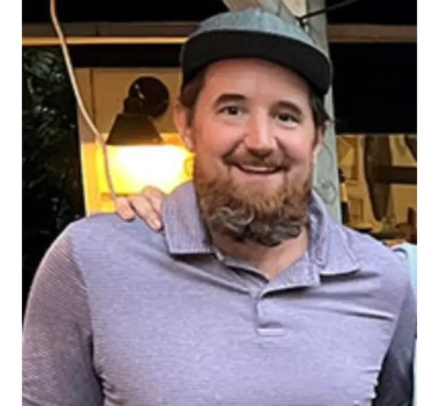 A smiling bearded man in a grey polo shirt and a flat cap, a lit lamp and a window frame behind him.