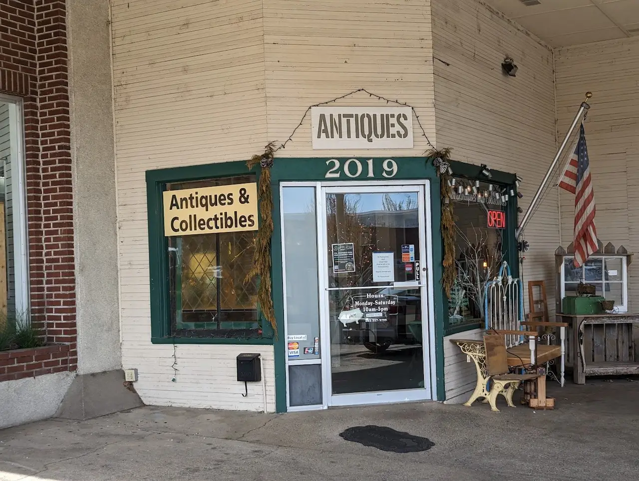 The corner storefront of an antiques shop in a white-painted clapboard building, a hand-lettered Antiques sign over the door and benches and cabinets out on the sidewalk.